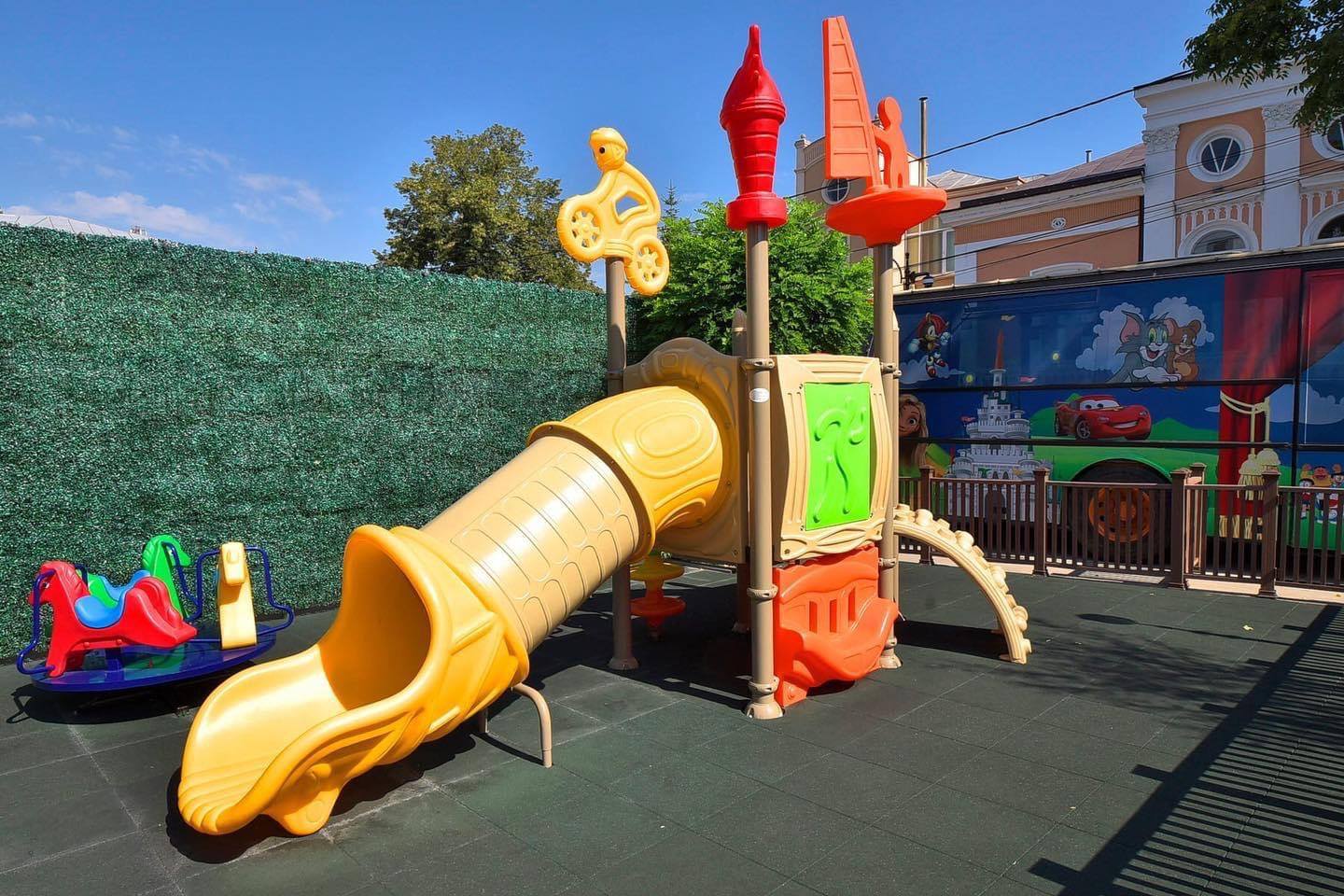Complex de joacă pentru exterior amplasat pe terasa unui restaurant family-friendly, cu tobogan, elemente de cățărare și suprafață de siguranță pentru copii.