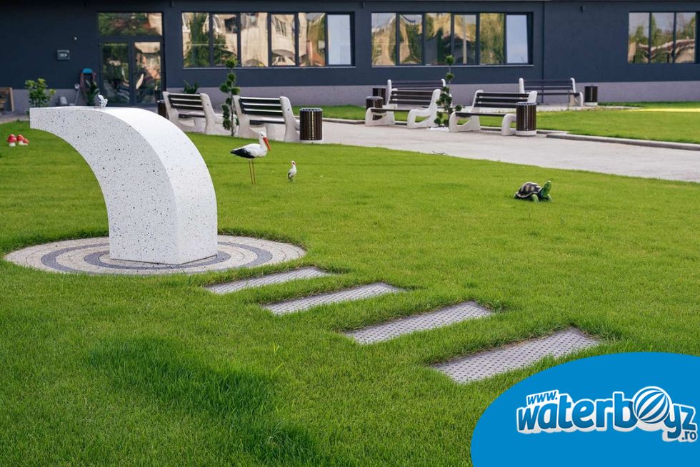 Cișmea urbană amplasată într-un parc rezidențial modern, cu spații verzi, alei pietonale și mobilier urban.