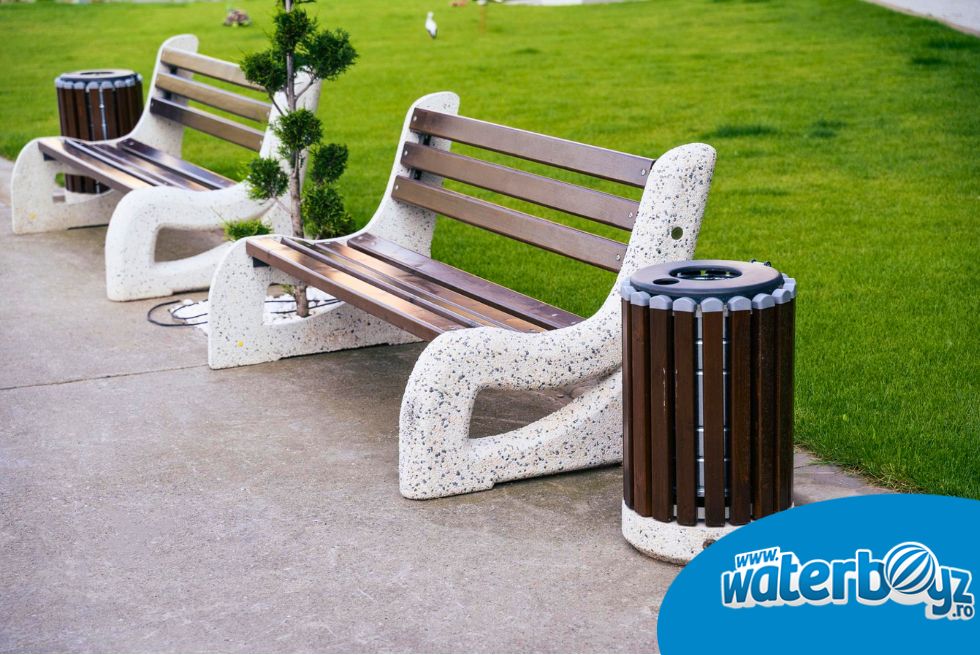 Bănci urbane și coșuri de gunoi amplasate într-un parc rezidențial modern cu spații verzi și alei pietonale.